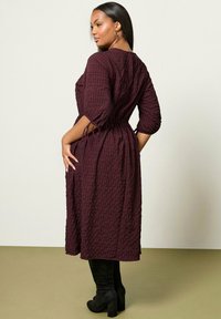 Woman wearing a burgundy checkered midi dress with 3/4 sleeves tied at cuffs, paired with black heeled knee-high boots, standing sideways.