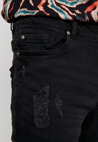 Denim negro con una textura desgastada, que presenta secciones deshilachadas y herrajes de plata. Se sitúa en la cintura con un diseño ajustado.