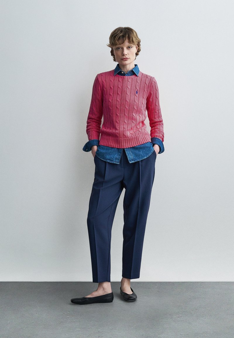 Personne debout les mains dans les poches, portant un pull rose à torsades sur une chemise en denim bleu, un pantalon bleu marine et des ballerines noires sur un fond uni.