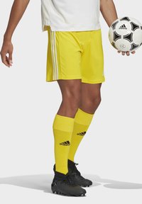 Gele voetbalkorte broek met drie witte strepen aan de zijkanten, gecombineerd met bijpassende gele sokken en zwarte voetbalschoenen, terwijl hij een zwart-wit bal vasthoudt.