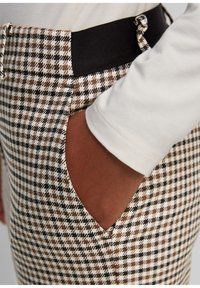 Pantalon à motif pied-de-poule marron et noir avec une ceinture élastique noire et une poche latérale ; porté avec une chemise à manches longues de couleur crème.