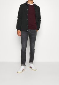 Man med svart knäppt jacka, vinröd skjorta, mörkgrå skinny jeans, vita strumpor och vita sneakers med röda och blå ränder som står och poserar.