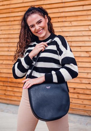 Black leather shoulder bag with a curved shape, featuring a subtle logo and a long strap. Model wearing a black and white striped sweater.