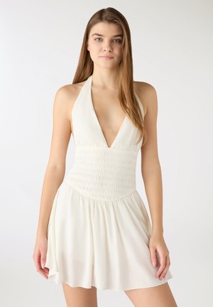 Giovane donna con capelli lunghi e lisci che indossa un vestito bianco senza maniche con scollo a V profondo e vita aderente arricciata.