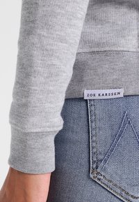 Dettaglio ravvicinato di un polsino e di un orlo di un maglione grigio con un'etichetta "Zoe Karssen" sopra la tasca posteriore di un paio di jeans in denim blu.