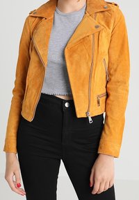 Gul mockajacka med asymmetrisk dragkedja och tryckknappar, i kombination med en grå kortärmad tröja och svarta högmidjade byxor.