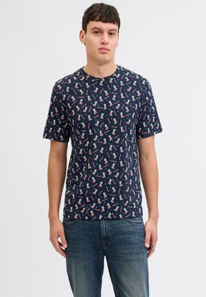 Jeune homme portant un t-shirt bleu marine avec un motif de canne en sucre d'orge et de chaussette de Noël, et un jean bleu, debout devant un fond clair uni.