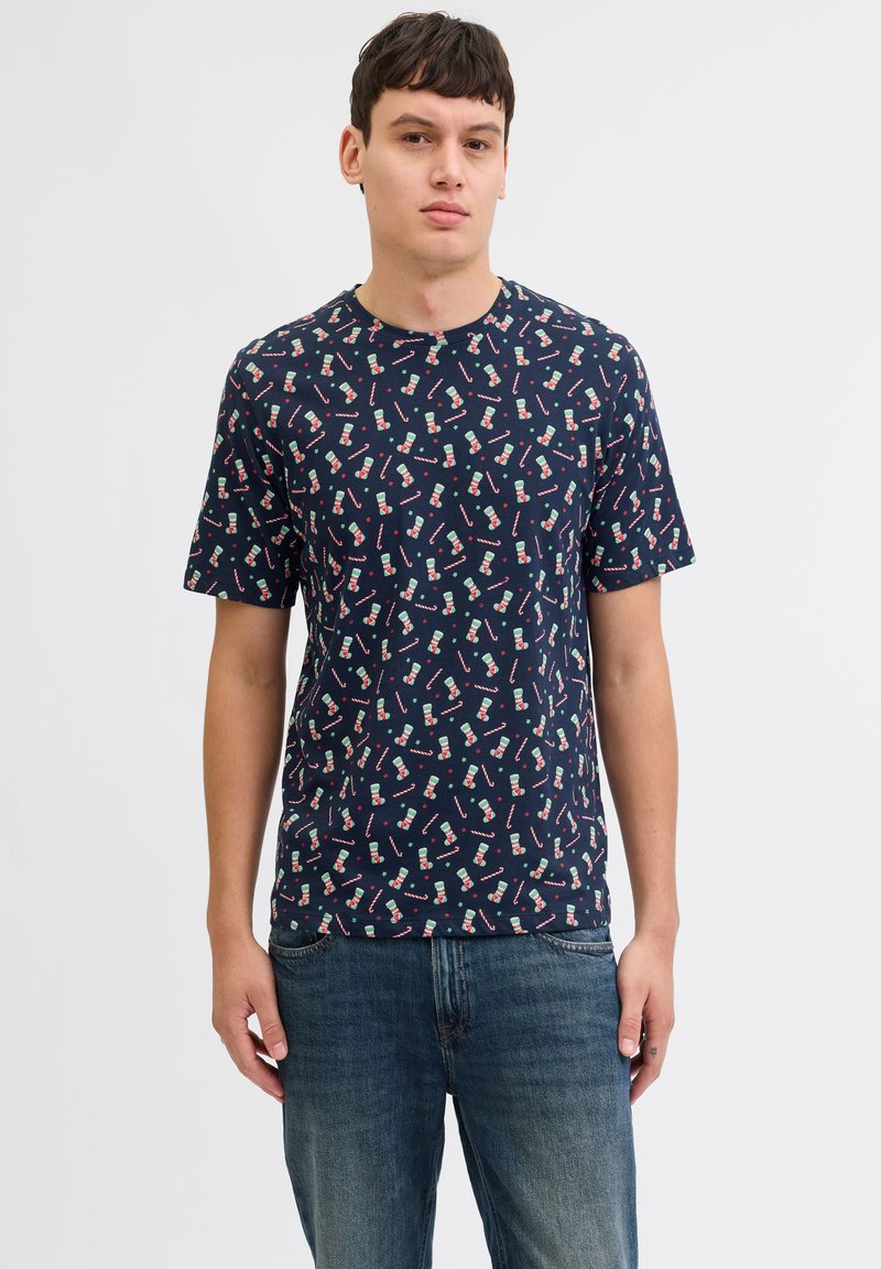 Junger Mann trägt dunkelblaues T-Shirt mit Zuckerstange- und Weihnachtssockenmuster und blaue Jeans, steht vor einem einfarbigen hellen Hintergrund.