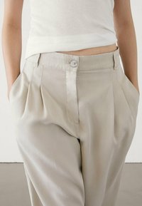 Pleated beige trousers with a smooth texture, featuring a front button closure and side pockets, paired with a white ribbed top.
