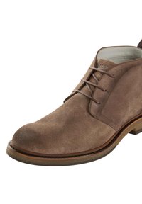 Stivale alla caviglia in suede marrone con punta arrotondata, dettagli cuciti, chiusura con lacci e suola in legno marrone chiaro. Fodera interna liscia.