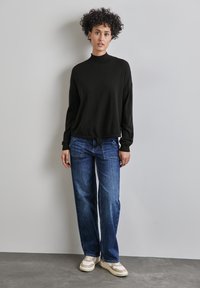 Schwarzer, langärmeliger Pullover mit hohem Kragen; kombiniert mit lässigen, verwaschenen blauen Jeans und beigen Sneakers mit weißen Akzenten.