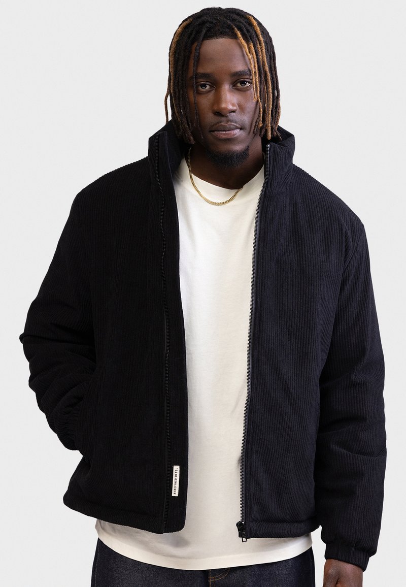 Άνδρας με dreadlocks μέχρι τους ώμους, φοράει μαύρο μπουφάν με φερμουάρ πάνω από λευκό πουκάμισο, στέκεται μπροστά σε μονόχρωμο φόντο.