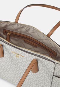 Geantă tote gri și bej cu un model de logo, accente din piele maro, două mânere superioare și închidere cu fermoar care dezvăluie un interior căptușit.