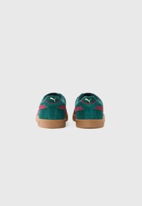 Sneakers din suede verzi și maronii cu talpă din cauciuc, văzute din spate, expunând logo-ul auriu Puma pe călcâie, pe un fundal alb simplu.