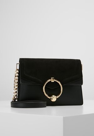 Bolso bandolera negro con cadena dorada y gran cierre circular dorado, exhibido sobre superficie blanca contra un fondo gris.