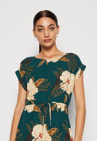 Vestido floral em verde-água com mangas curtas. Apresenta uma faixa na cintura, flores em creme e castanho, e padrões de folhagem verde. Tecido suave e leve.