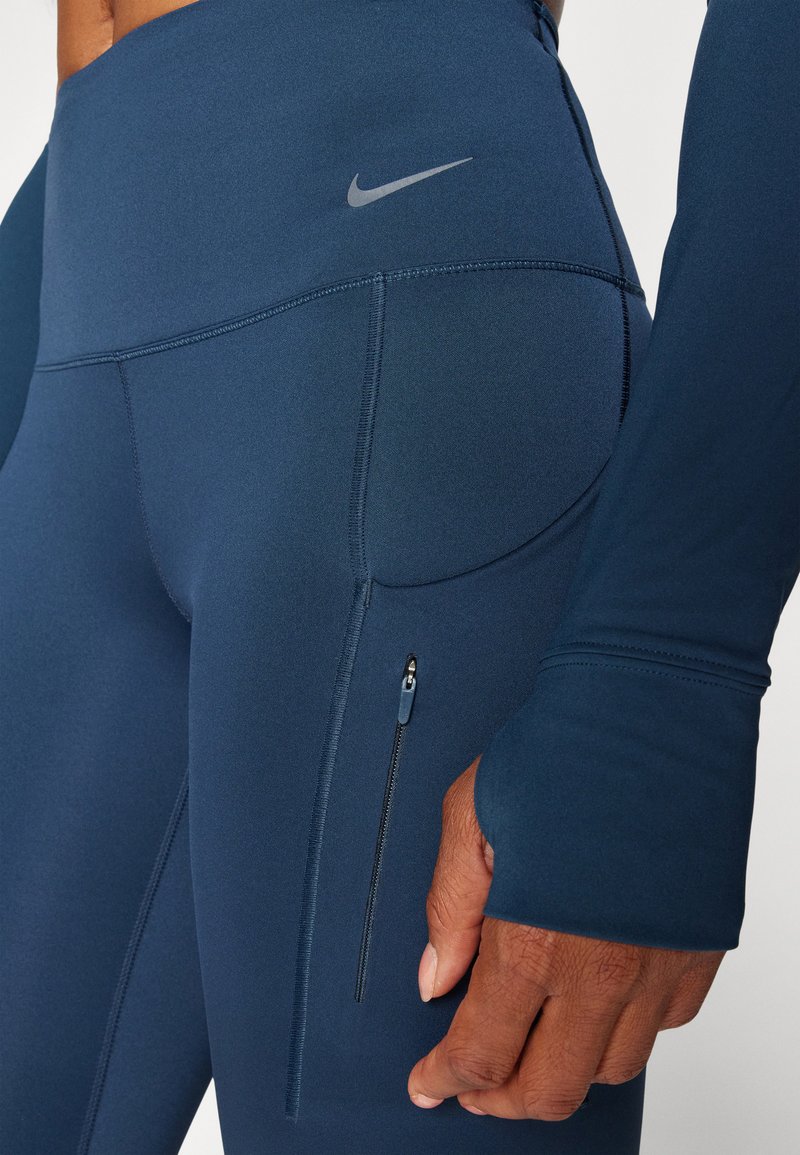Granatowe legginsy sportowe z wysokim stanem, gładki materiał, boczna kieszonka na zamek oraz małe szare logo Nike na pasie. 