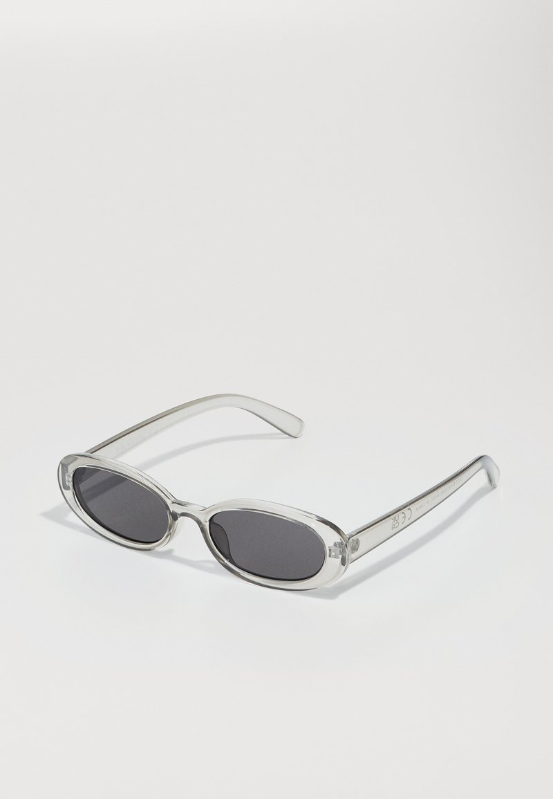 Gafas de sol con forma ovalada, montura de plástico transparente, lentes gris oscuro y patillas delgadas y transparentes con acentos de logotipo y un diseño minimalista.