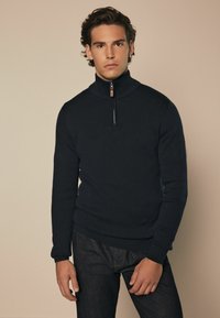 Marinekleurige zip-neksweater van een gladde breimateriaal met een hoge kraag, voorzien van een zilveren rits en geribbelde manchetten, gecombineerd met donkere spijkerbroeken.