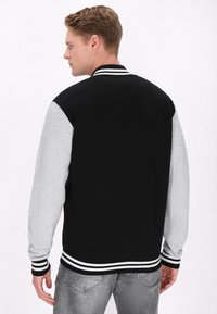 Jachetă de tip varsity neagră și gri, cu guler ribbit și manșete cu dungi, având spatele solid și mânecile în contrast. Țesătură texturată.
