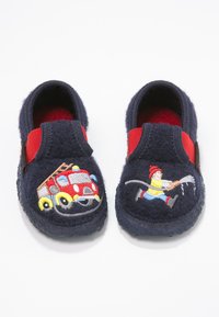 Chaussons pour enfants bleu marine avec côtés élastiques rouges, décorés d'un camion de pompiers brodé à gauche et d'un pompier aspergeant de l'eau à droite.