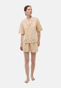 Ensemble de pyjama en tissu pêche doux, composé d'une chemise à manches courtes avec poches avant et des détails brodés blancs, associé à un short assorti.