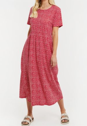 Kobieta w czerwonej sukience midi w białe kropki, z krótkimi rękawami, okrągłym dekoltem oraz beżowymi sandałami na klamrę, stojąca na białym tle.