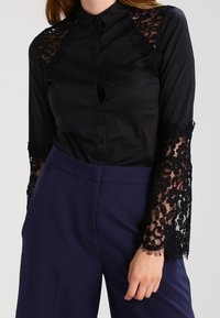 Chemise noire à boutons avec des manches longues en dentelle et des bords festonnés. Tissu lisse avec des accents en dentelle transparente sur les épaules et les poignets.