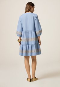 Vestido azul claro con mangas abullonadas, dobladillo de volantes en capas y un patrón geométrico bordado en oro a lo largo del fondo y en los puños.