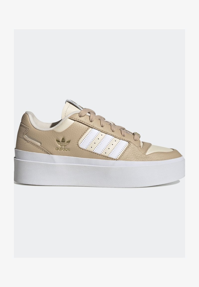 Beige ja cream Adidas-sneaker, jossa on valkoiset raidat, kullanvärinen logo sivussa, beige nauhat ja paksu valkoinen platform-pohja, sivuprofiili valkoisella taustalla.