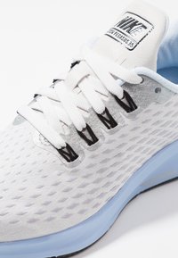 Scarpa sportiva bianca con tomaia in rete, motivo testurizzato, dettagli argento, lacci neri e suola azzurro chiaro. Presenta un'etichetta con il marchio sulla linguetta.