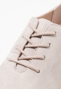 Zapato de ante beige claro con puntera redonda, que presenta cordones beige y una superficie texturizada. El diseño incluye mínimos acentos de costura.