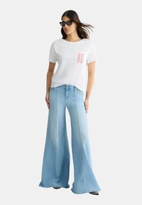 T-shirt bianca a maniche corte con stampa del logo rossa, abbinata a jeans a zampa d'elefante di colore blu chiaro. I jeans hanno una texture sbiadita e cuciture classiche.