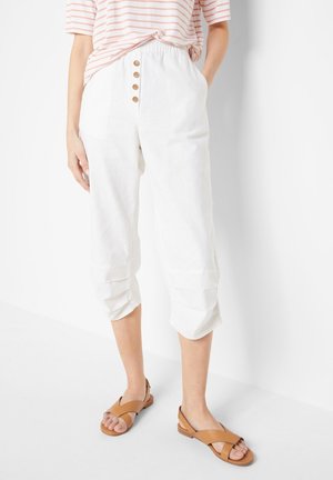 Femme portant un pantalon blanc court avec des détails de boutons et des sandales plates couleur camel, debout avec une main dans la poche sur un fond blanc.
