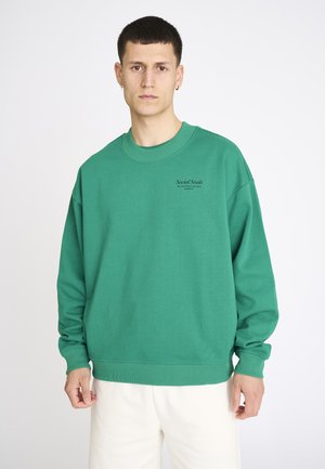 Ung mand med kort hår iført en grøn sweatshirt med rund hals med teksten "Social Souls" og hvide bukser mod en ensfarvet baggrund.
