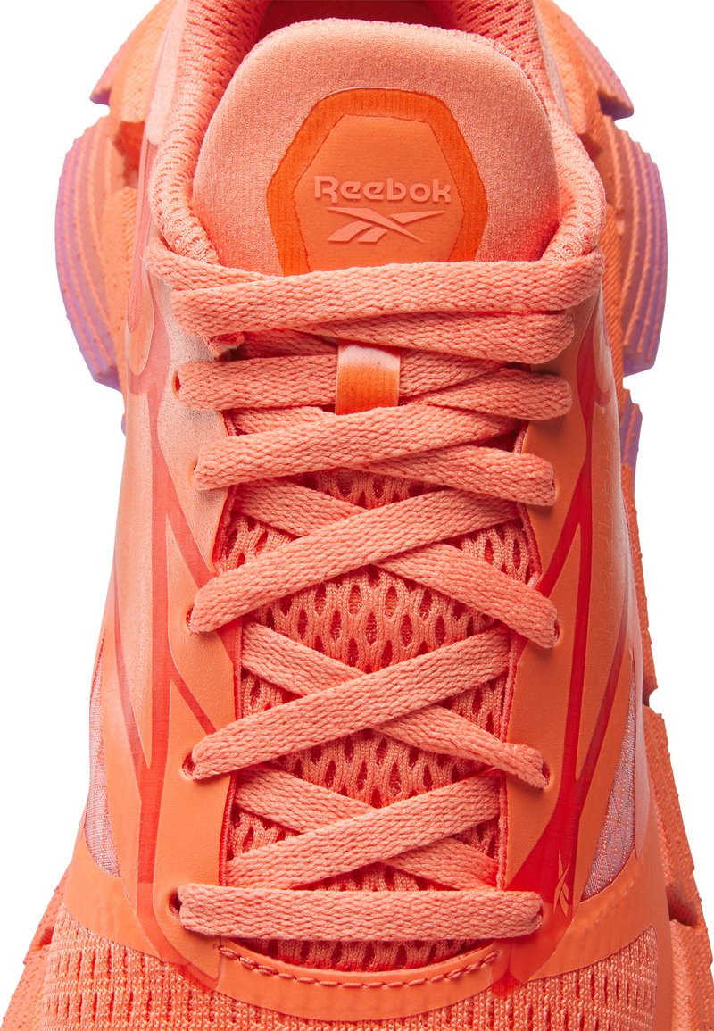 Sneaker Reebok con una toma superiore in rete arancione brillante, lacci piatti e una suola in gomma testurizzata. Presenta un logo esagonale sul tallone.