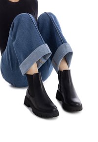 Botas de cuero negras hasta el tobillo con tacones bajos y gruesos, que cuentan con paneles laterales elásticos y suelas de goma texturizada. Combinadas con pantalones de denim con vuelta.