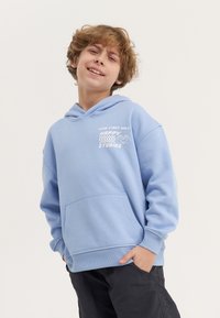 Gaiši zils kokvilnas kapuce ar kangaroo kabatu, kapuci un baltu grafisku tekstu "Labas vibrācijas tikai Happy Studios" uz priekšpuses.