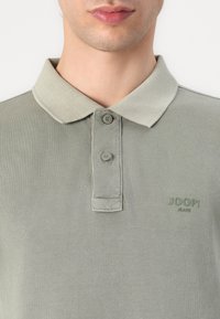 Tricou polo gri, dintr-un material texturat, cu deschidere cu două butoane și guler. Logo-ul mărcii "JOOP!" este brodat în verde asortat.