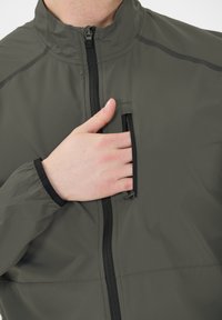 Person som bär en grå jacka med en svart dragkedja, som stoppar sin hand i en bröstficka med vertikal dragkedja.