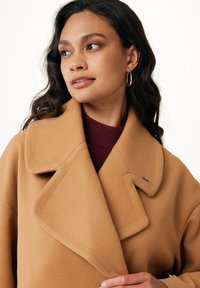 Cappotto di lana color cammello con ampio colletto e grandi revers. Presenta un piccolo dettaglio metallico ed è indossato sopra un maglione bordeaux.