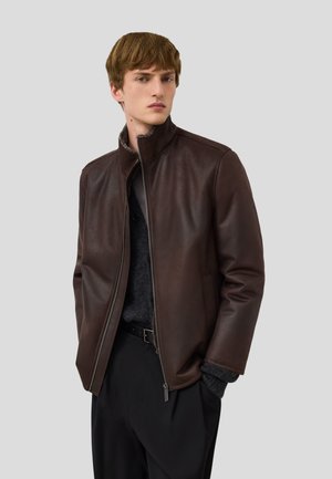 Jeune homme portant une veste en cuir marron foncé sur un pull noir et un pantalon noir, se tenant devant un fond gris clair uni.