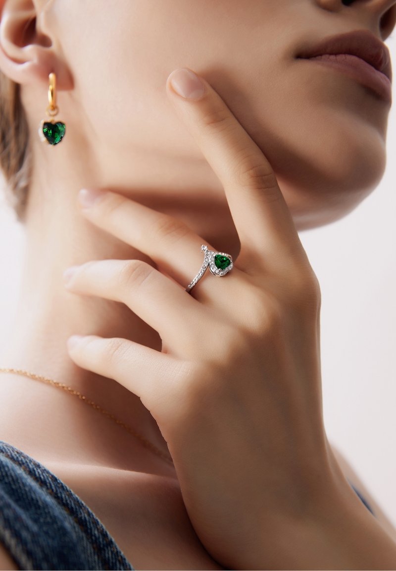 Gros plan sur une main touchant la ligne de la mâchoire, portant une bague en argent avec une pierre précieuse verte en forme de cœur et une boucle d'oreille assortie en forme de cœur vert.