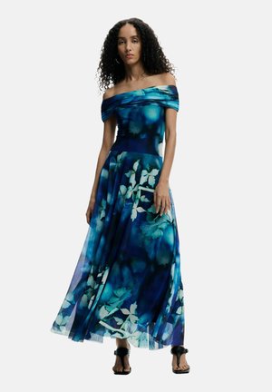 Mujer con pelo rizado, con un vestido maxi azul con flores y hombros descubiertos y tacones negros abiertos, de pie frente a un fondo liso.