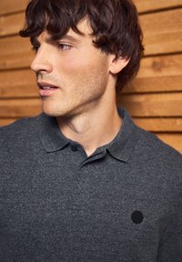 Mladý muž s tmavě hnědými vlasy, oblečený v šedém svetr s límečkem, dívající se vlevo na pozadí dřevěných latěk.