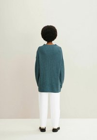 Pull en maille bleu sarcelle à coupe décontractée, manches longues et texture côtelée, associé à un pantalon blanc à jambes larges et des chaussures noires.