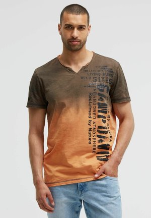 Man with short hair and beard wearing brown to orange gradient V-neck T-shirt with vertical black text and light blue jeans, standing against plain background.