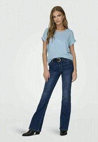 Ung kvinne står med hånden hvilende på låret iført en lyseblå kortermet topp, mørkeblå vide jeans, svart belte og svarte ankelstøvler.