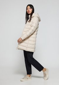 Abrigo tipo plumas beige con capucha, diseño acolchado, cierre de cremallera y bolsillos laterales, combinado con jeans oscuros y zapatillas de color beige claro.