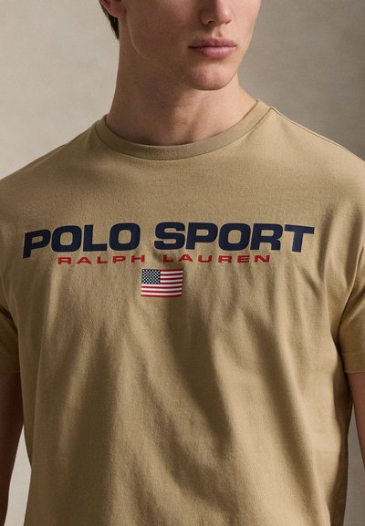 Μπεζ βαμβακερό μπλουζάκι με την επιγραφή "POLO SPORT" σε ναυτικό μπλε και "RALPH LAUREN" σε κόκκινο, με ένα μικρό γραφικό αμερικανικής σημαίας. Κοντά μανίκια, στρογγυλή λαιμόκοψη.